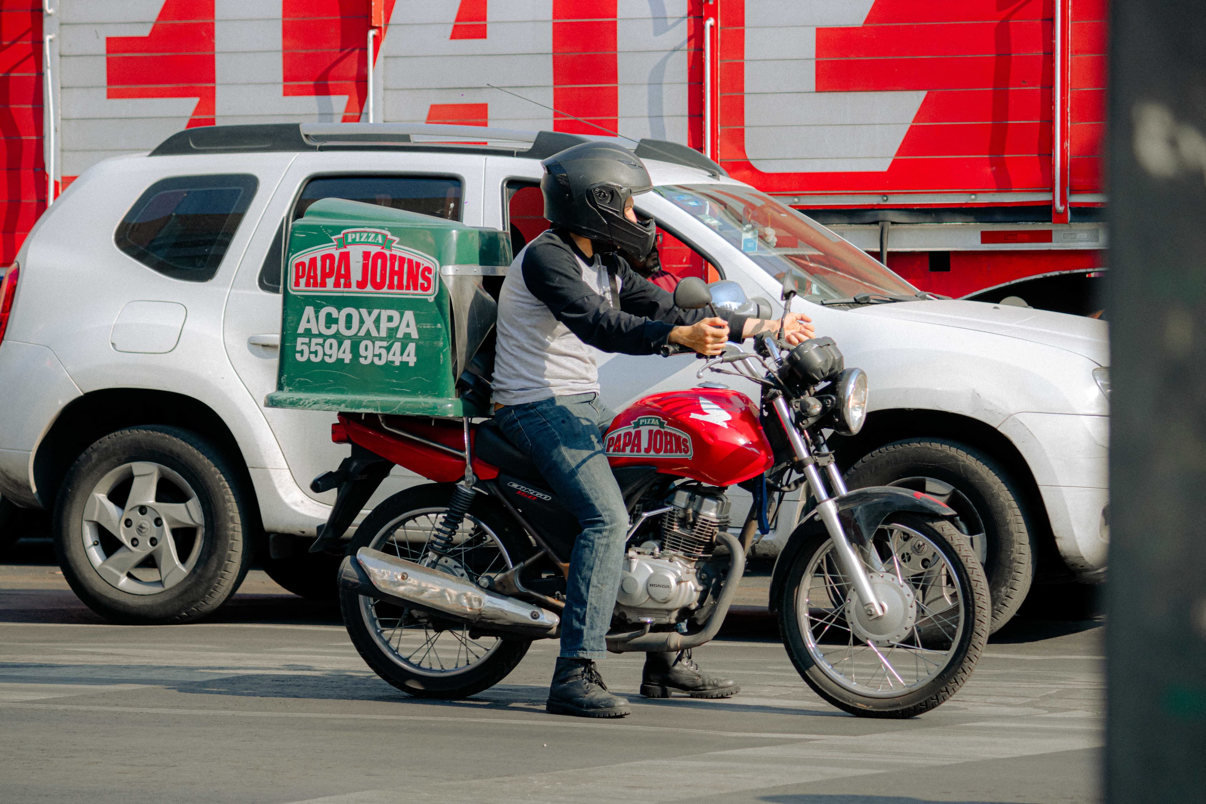A pizza delivery rider on a motorcycle waiting at a busy city street intersection.