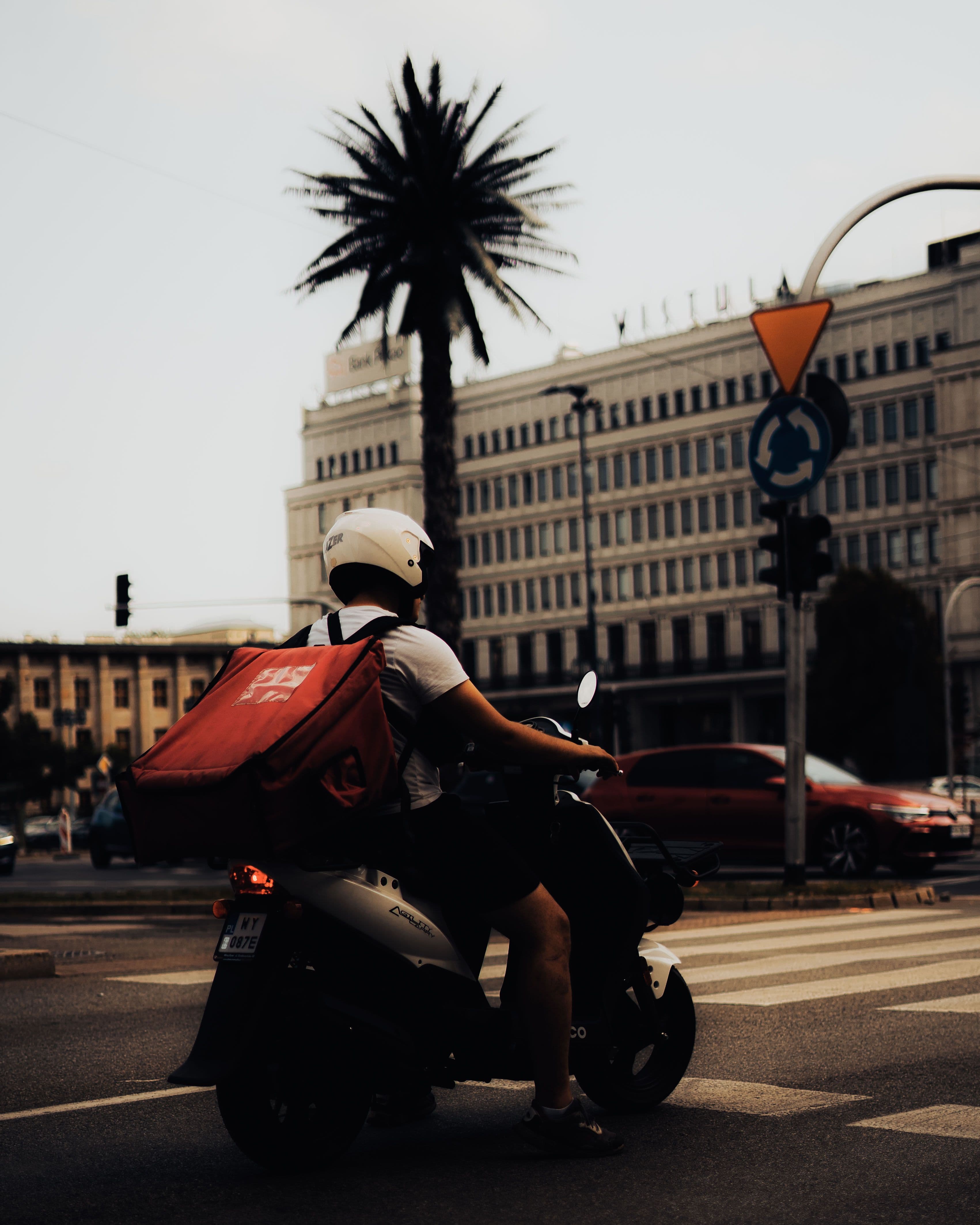 A delivery rider on a motorcycle wearing a helmet, riding through a city street with buildings in the background.