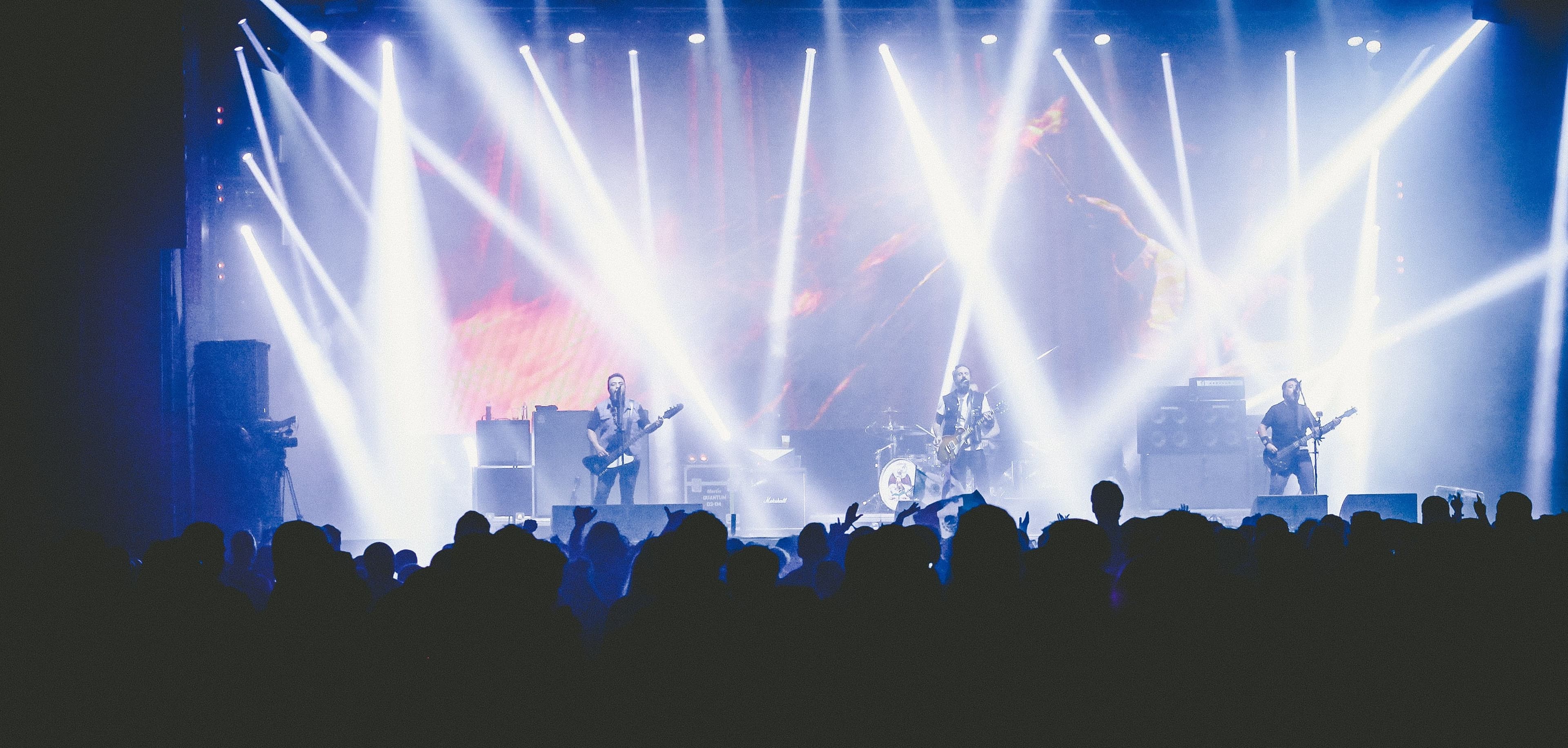 A band performs on a smoke-filled stage under bright concert lights as a crowd watches.
