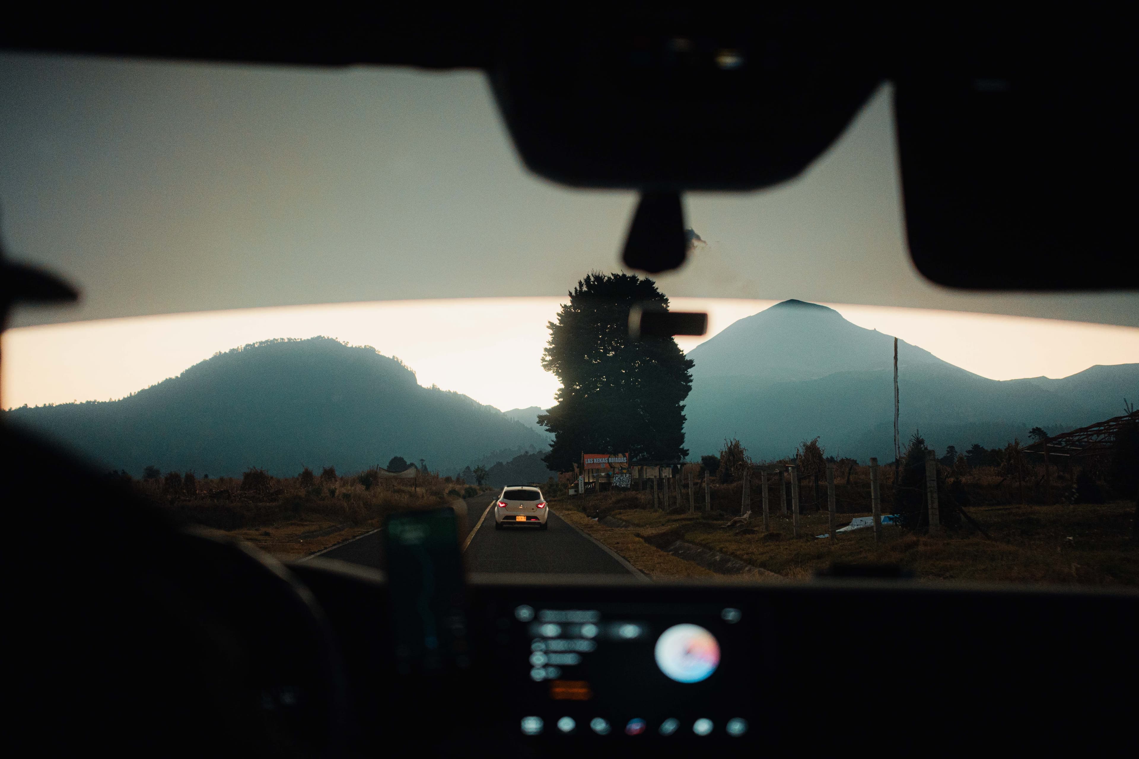 A car drives along a scenic mountain road at sunset, seen from inside the vehicle looking out through the windshield.