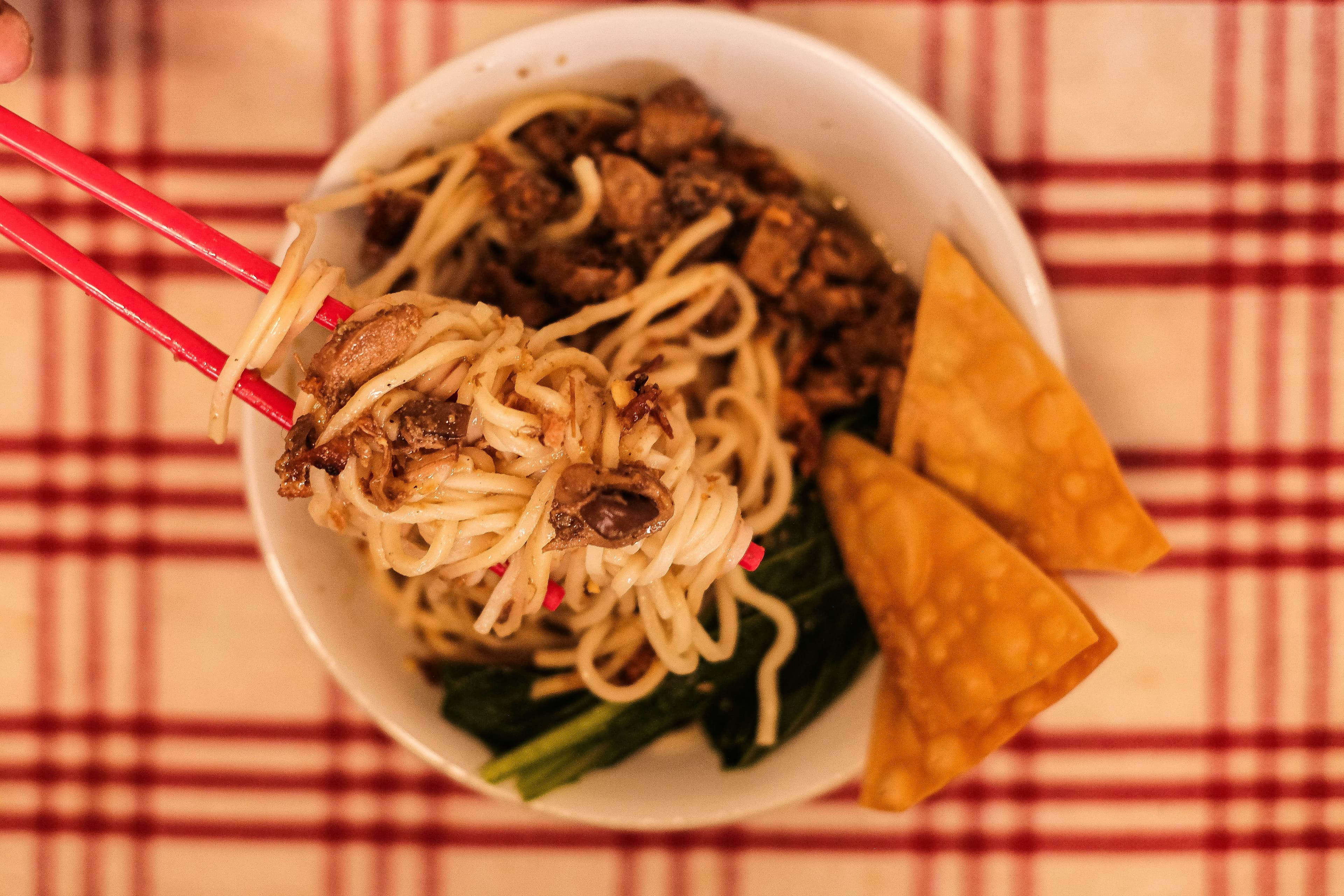 A bowl of Asian egg noodles topped with crispy wontons, minced meat, and vegetables, photographed from above with a plaid tablecloth in the background.