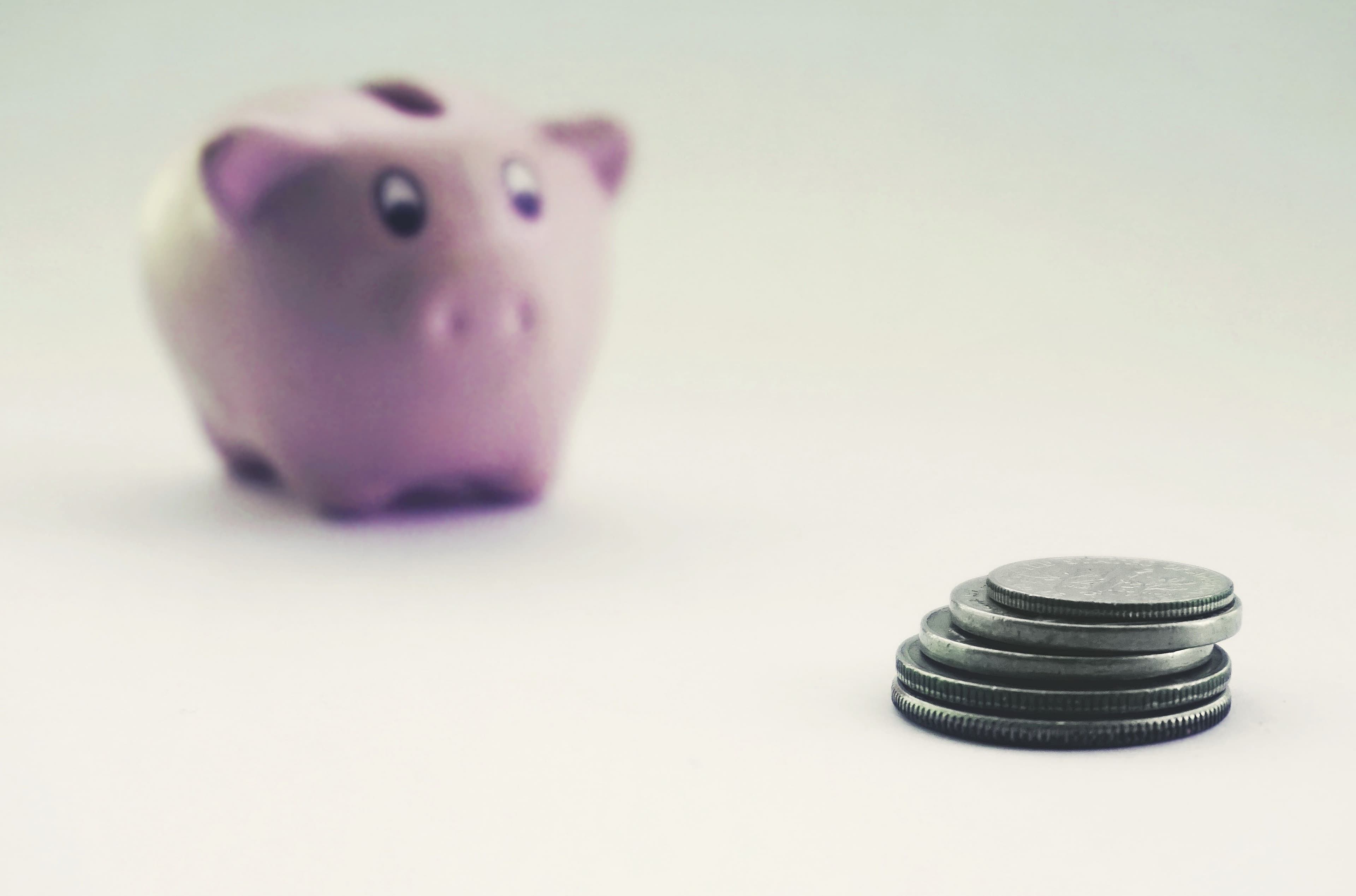 A pink piggy bank blurred in the background behind a neat stack of coins on a plain white surface.