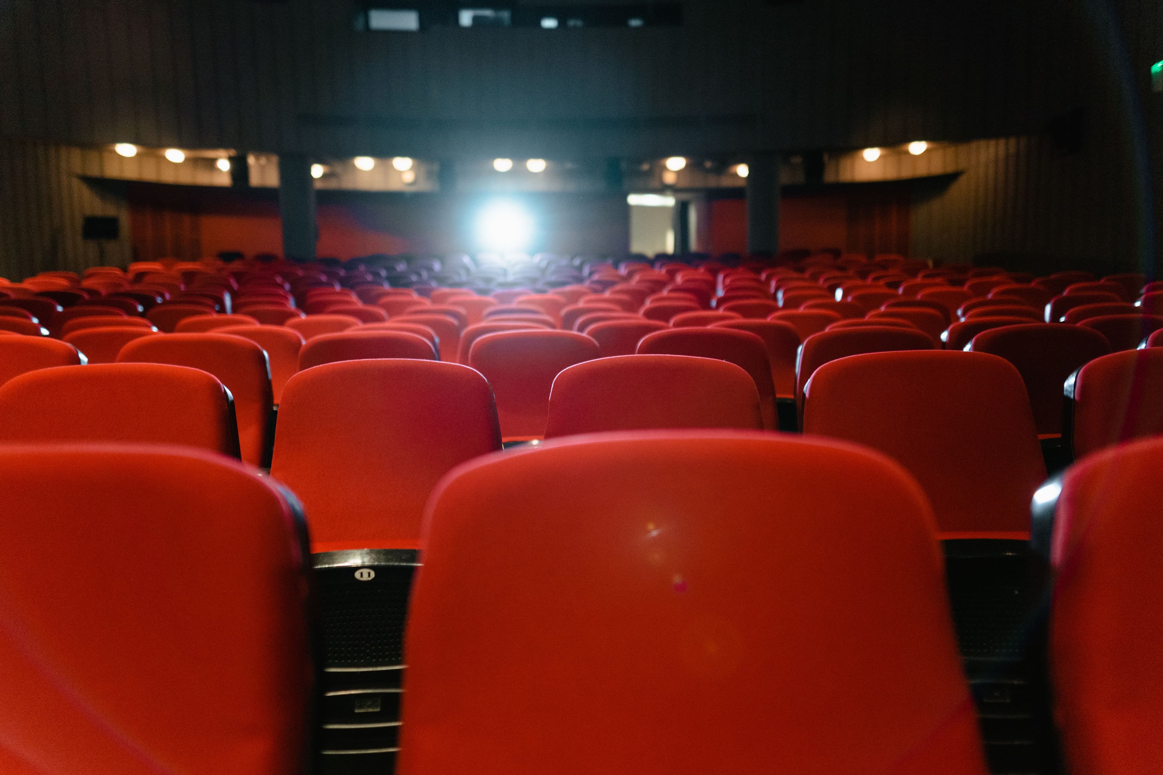 Rows of empty red cinema seats facing an illuminated screen in a darkened movie theatre.