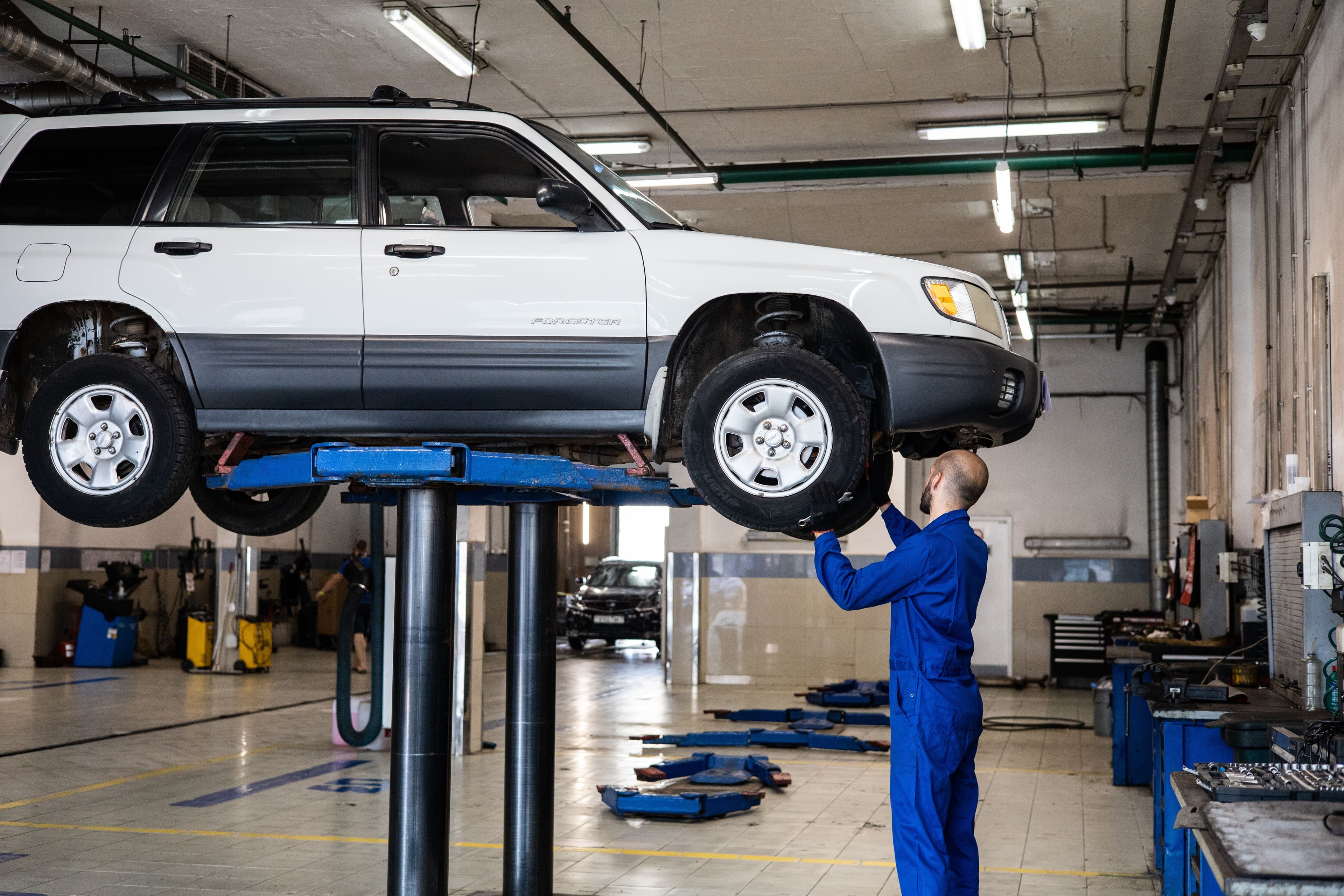 A mechanic in blue coveralls inspecting a white SUV lifted on a hoist inside a well-lit auto repair garage.