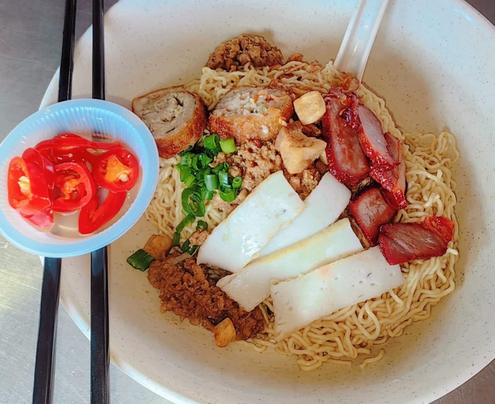 A bowl of Sarawak kolo mee topped with char siew, minced pork, fish cake slices and spring onion, with sliced chilli in vinegar on the side