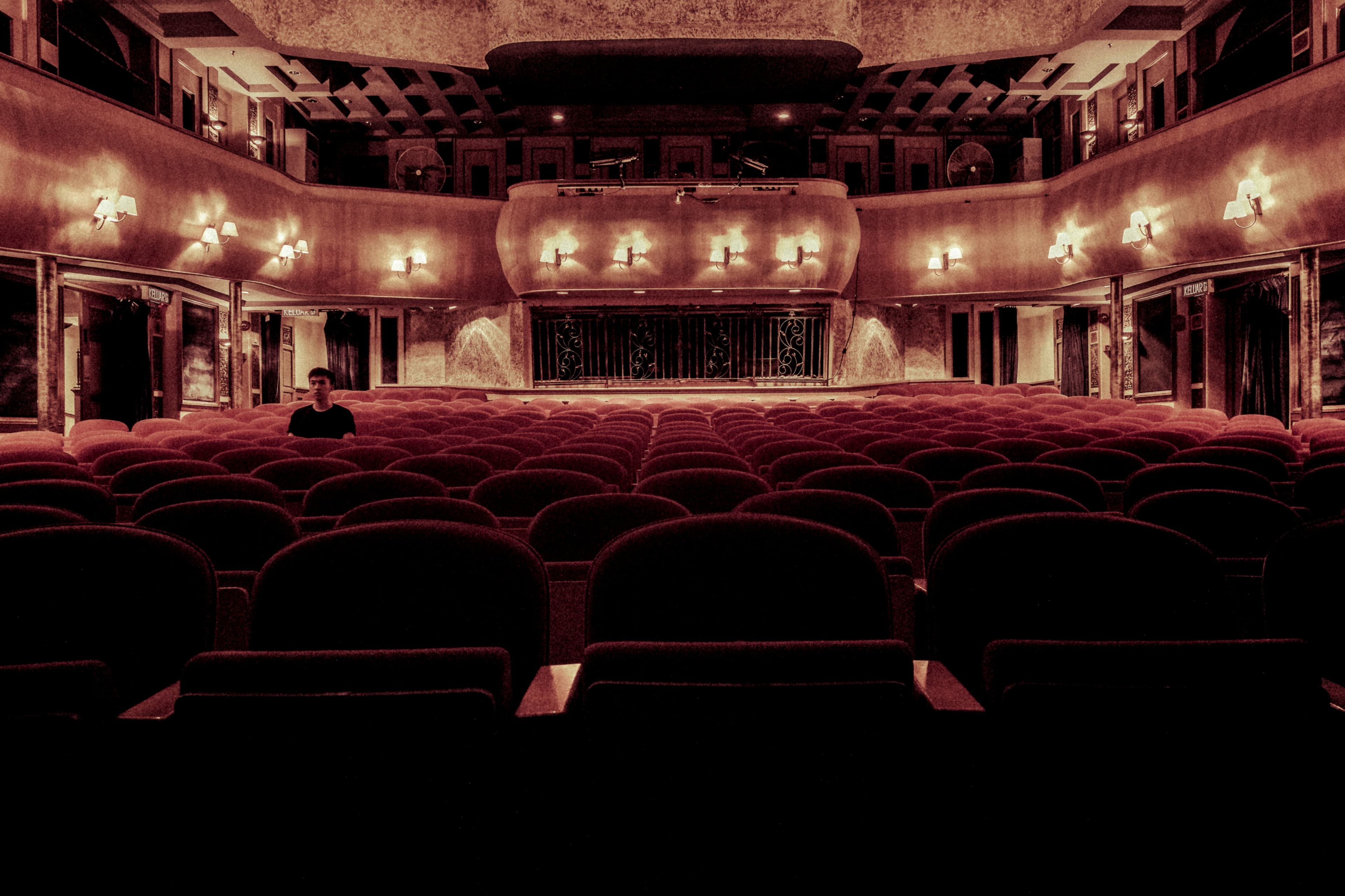 A wide, empty vintage cinema auditorium with rows of red velvet seats and warm ambient lighting facing the screen.