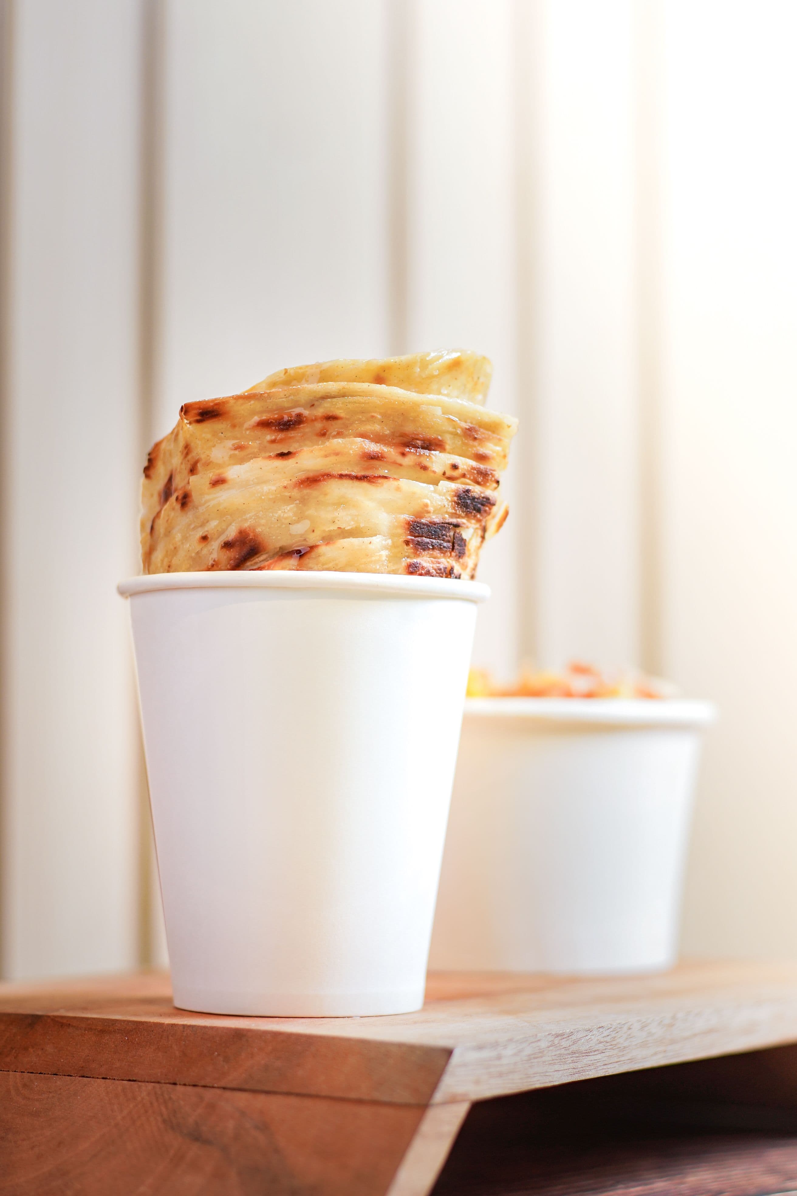 Crispy, flaky layers of golden-brown roti canai stacked upright in a white cup on a wooden surface, showing off the bread's texture.