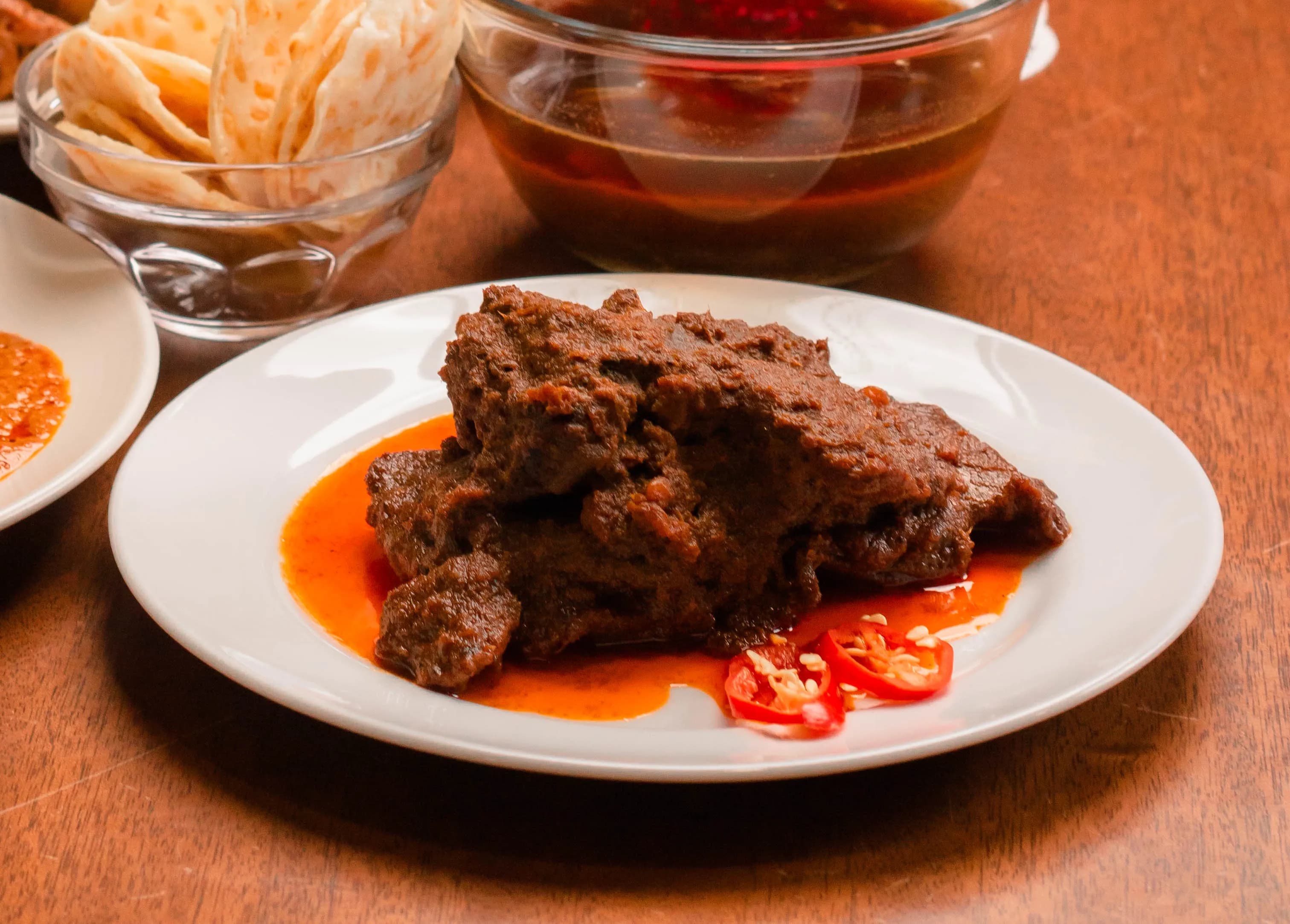 Close-up of a plate of beef rendang with chilli and sides on a wooden table.