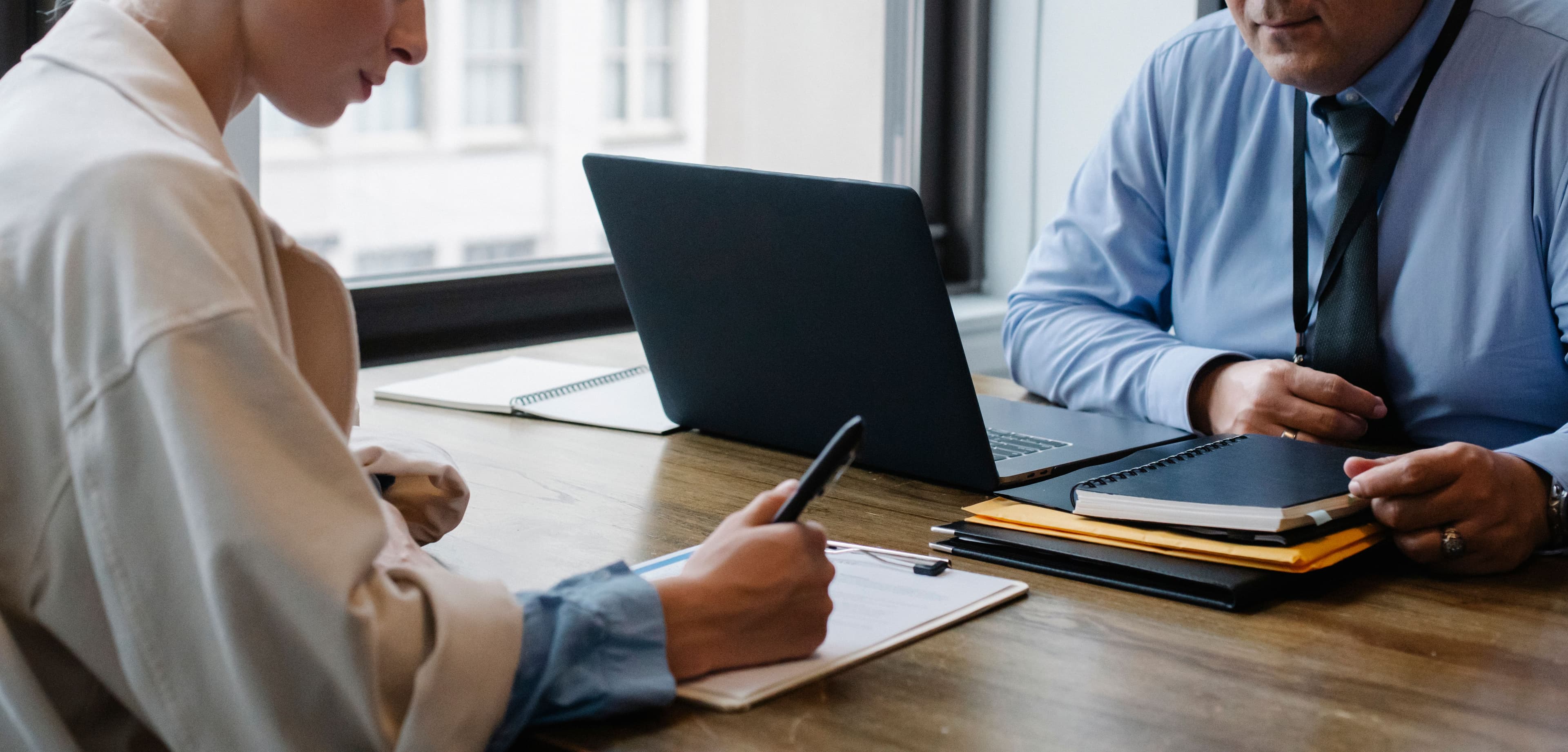 Two office workers sit at a desk reviewing printed documents and a laptop while signing paperwork together.