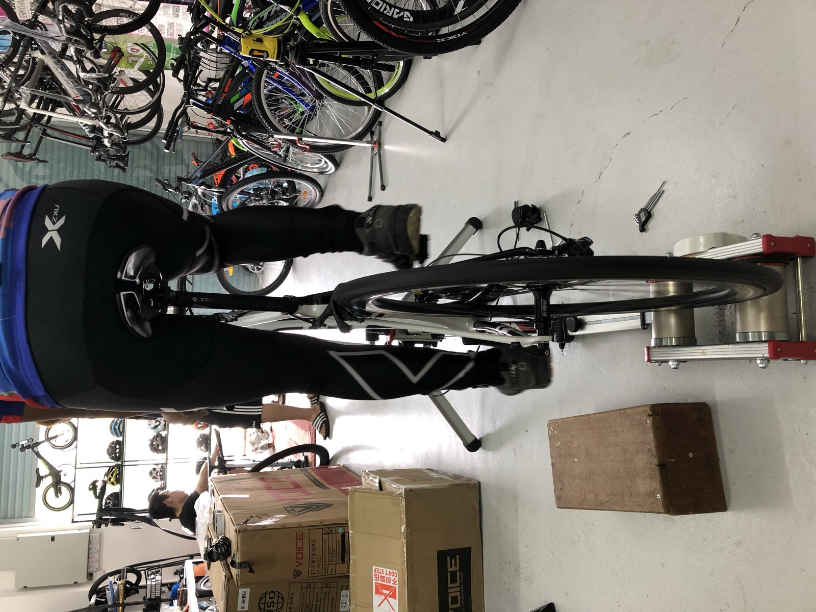 A cyclist riding a road bike mounted on an indoor turbo trainer inside DC Cycles in Seremban, with bicycles and helmets lining the shop walls behind